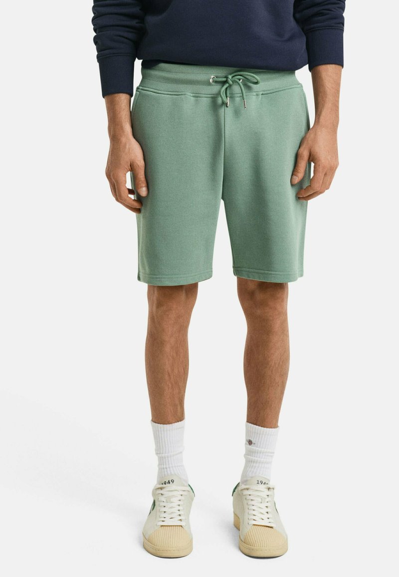 Pantalones cortos de algodón verde claro con cintura elástica y cordón. Combinados con calcetines blancos y zapatillas beige. Diseño simple y casual.