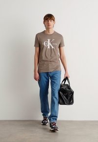 Rudas medvilninis marškinėliai su baltu Calvin Klein logotipu, mėlyni džinsai ir juoda odinė rankinė. Sportbačiai turi daugiashuolius akcentus.