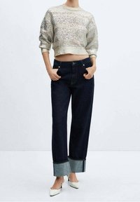 Suéter texturizado color beige claro con detalles de brillo, combinado con jeans de pierna ancha azul oscuro con puños de denim doblados y tacones blancos.