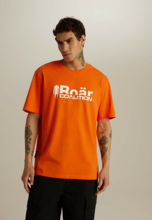 Pomarańczowa bawełniana koszulka z krótkim rękawem, z dużym białym nadrukiem, na którym widnieje napis "Roär COALITION". Model nosi czarne spodnie cargo.
