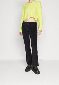 Un pull cropped côtelé vert lime associé à un pantalon en velours côtelé noir à pattes d'eph, avec une taille à boutons et des chaussures à plateforme noires.