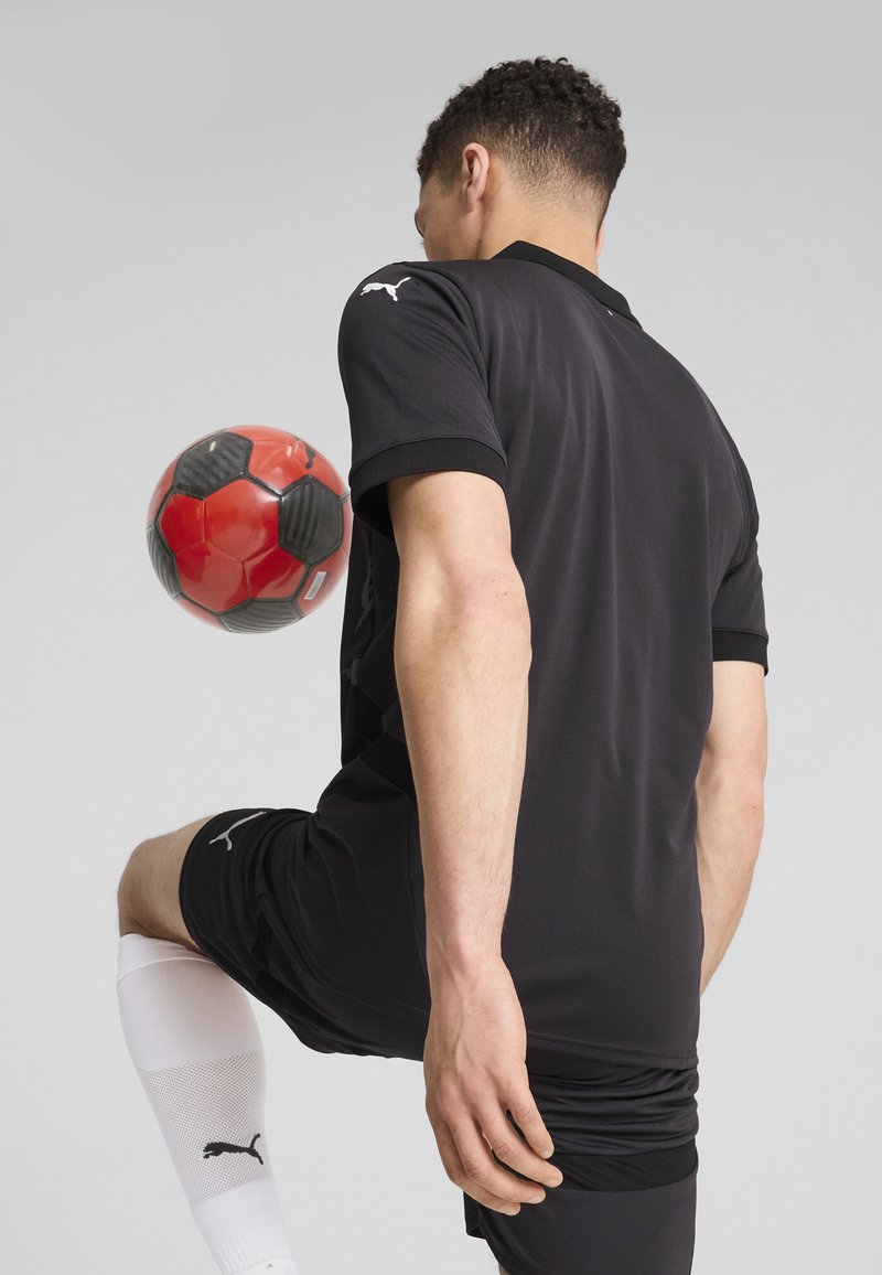Jersey atlético negro y pantalones cortos con acentos negros. El jugador equilibra una pelota de fútbol roja y negra en su rodilla, vistiendo calcetines blancos.