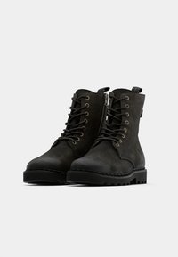 Botines de cuero negro con cierre de cordones y cremallera. Suelas gruesas de goma con acabado texturizado y tacón reforzado, con ojales metálicos.