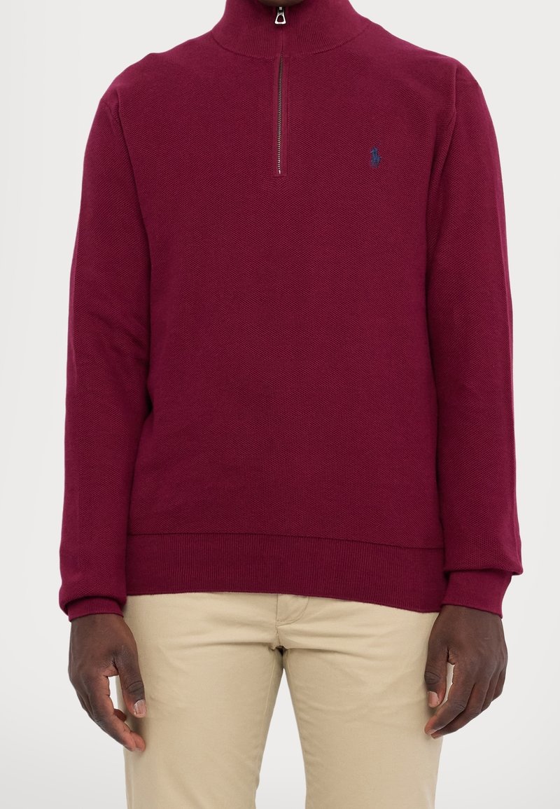 Homme portant un pull bordeaux à col zippé avec poignets et ourlet côtelés, associé à un pantalon beige, debout devant un fond neutre.
