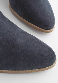 Pantofi din suede bleumarin cu vârf ascuțit și talpă din lemn. Prezintă un detaliu cusut în jurul marginii pentru un aspect suplimentar.