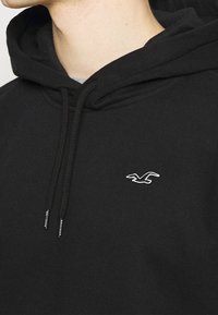 Primer plano de una persona que lleva una sudadera negra con cordones y un pequeño logo de gaviota blanca en el pecho.
