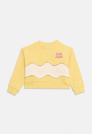 Kollane lühike pikkade varrukatega dressipluus lainelise valge triibuga rinna peal ja roosa tekstiga "Club Soleil" vasakul ülaosas.