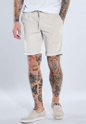 Mann trägt hellbeige Shorts und Slip-On-Schuhe, zeigt tätowierte Beine und Arme mit Motiven wie einer Rose, einem Schmetterling, Sonne und Schriftzug.