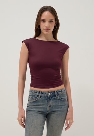 Donna con lunghi capelli castani che indossa orecchini a cerchio dorati, un top senza maniche aderente viola scuro e jeans blu a vita alta su uno sfondo semplice.