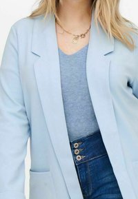 Light blue blazer with notched lapels, textured fabric, and side pockets, layered over a blue top, paired with high-waisted dark denim.