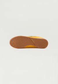 Gelbe Sneaker mit einem strukturierten Wildleder-Obermaterial, schwarzen Akzenten und einer braunen Gummisohle mit kreisförmigen Profilmustern. Flaches, niedriges Design.