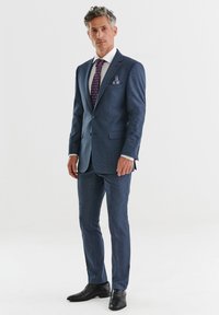 Granatowy garnitur z fakturowanej tkaniny, zapięcie na jeden guzik, biała koszula, krawat w wzory oraz chusteczka w kieszeni; czarne buty dopełniają cały wygląd.