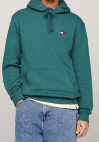 Felpa teal con cappuccio e coulisse, tasca frontale e un piccolo patch con logo nei colori rosso, bianco e blu sul lato sinistro del petto. Jeans in denim visibili.