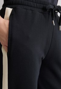 Nahaufnahme von schwarzen Sweatpants mit beigen Seitenstreifen, elastischem Bund mit Kordelzug und einer Hand, die in der vorderen Tasche ruht.