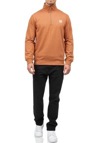 Homme portant un pull à fermeture éclair quart orange rouille, pantalon noir et baskets blanches, debout devant un fond blanc.