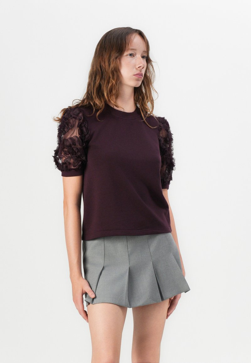 Camicia a maniche corte di colore viola scuro con dettagli in tessuto floreale trasparente sulle maniche, abbinata a una mini gonna a pieghe grigia.