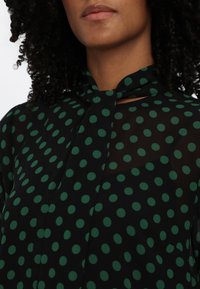 Gros plan d'une personne portant un chemisier noir transparent avec de grands pois verts et un détail noué au niveau du col.
