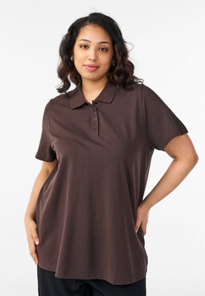 Kvinde med skulderlangt, bølget hår iført en løs, mørkebrun kortærmet polo, stående med den ene hånd på hoften.