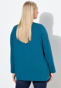 Blonde Frau mit glattem Haar trägt ein türkisfarbenes Langarmshirt und dunkle Jeans, steht seitlich gegen eine weiße Wand.