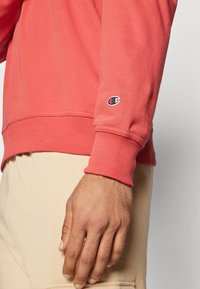 Koral genser med ribbestrikkede mansjetter, prydet med en liten svart og hvit logo på ermet. Materialet har en myk, strukturert overflate.