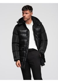 Hombre con chaqueta de plumas negra con capucha, camisa polo blanca y pantalones negros, mirando hacia un lado frente a un fondo liso.