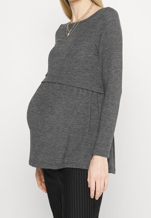 Zwangere vrouw draagt een grijs longsleeve zwangerschaps top en zwarte krijtstreeppantalon, met laagjes gouden kettingen.