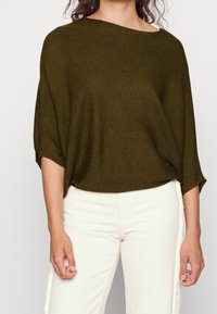 Olijfgroen gebreid sweater met een ruime bootnek, een losse pasvorm en een textuur van ribbels, gecombineerd met witte jeans met een raffelige zoom.