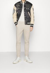 Veste bomber en satin noir avec manches crème, ornée de patchs et d'un design typographique. Pantalon beige ajusté et bottines blanches à lacets.