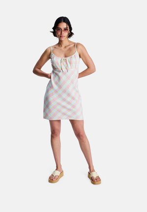 Vrouw draagt een pastelgeruite mouwloze jurk en beige platformsandalen, staat met handen in de zij tegen een witte achtergrond.