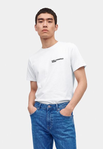 Jeune homme portant un t-shirt blanc Karl Lagerfeld Jeans et un jean bleu, les mains dans les poches sur un fond uni.
