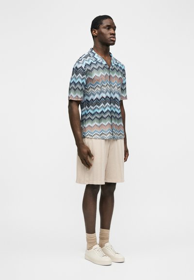 Άνδρας που στέκεται πλάγια φορώντας πουκάμισο με κοντά μανίκια και ζιγκ-ζαγκ μοτίβο, μπεζ σορτς, μπεζ κάλτσες και λευκά αθλητικά παπούτσια σε απλό φόντο.