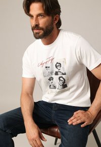 Hombre con barba que lleva una camiseta blanca con cuatro esqueletos en gafas de sol y sombreros, sentado en una silla marrón contra un fondo neutro.