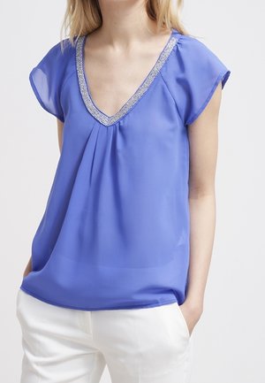 Frau trägt eine lockere, kurzärmelige blaue Bluse mit perlenverziertem V-Ausschnitt und weiße Hose, Hände in den Taschen, vor einfachem Hintergrund.