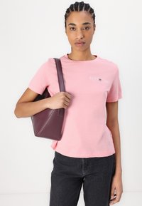 Lyserød bomulds t-shirt med korte ærmer, der har et lille hvidt logo-design. Sammen med en mørkegrå højtaljet nederdel og en bordeaux taske.