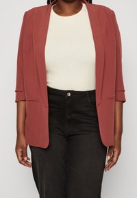 Ribbad vit topp under en roströd blazer med trekvartsärmar, som har en slät textur och öppen frontdesign, kombinerad med svarta byxor.