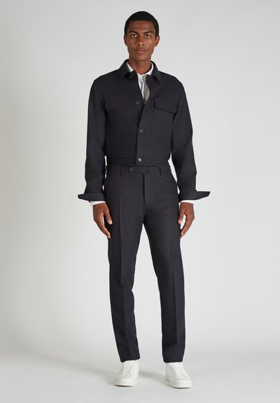 Giacca blu scuro con due tasche frontali, colletto con bottoni, abbinata a pantaloni blu scuro su misura e sneakers bianche. Finitura in tessuto testurizzato.