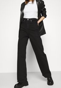 Jeans negros de cintura alta con piernas anchas, tela de denim texturizada, bolsillos frontales y cierre de botón. Combinados con una camiseta blanca y una blazer negra.