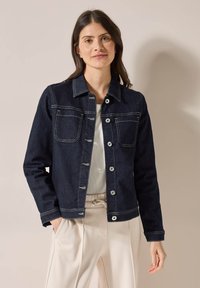 Veste en denim foncé avec deux poches poitrine, boutons en argent et couture blanche. Portée sur un haut clair et associée à un pantalon clair.