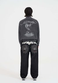 Person trägt einen dunklen Strickpullover mit "Ed Hardy" und Skull-Design sowie schwarze Hosen mit Spinnennetz-Stickerei auf den hinteren Taschen.