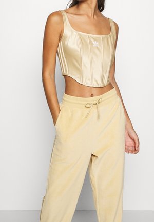 Femme portant un débardeur court beige brillant Adidas et un pantalon ample beige à cordon avec les mains dans les poches, sur un fond uni.