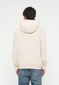 Mężczyzna z ciemnymi włosami, ubrany w beżowy hoodie i niebieskie dżinsy, odwrócony plecami na tle białego tła.