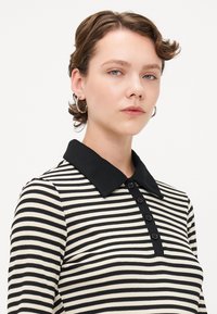 Svart og kremstripete langermet polo med svart ribbestrikket krage og fem knapper. Glatt tekstur og tettsittende design.