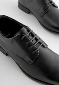Czarne skórzane buty do garnituru o gładkiej teksturze, z zapięciem na sznurowadła, okrągłym noskiem i niskim obcasem. Posiadają subtelne przeszycia i kontrastujące sznurowadła.