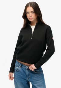 Zwarte gebreide trui met hoge kraag en halve rits; ribbelboorden en -zoom; gedragen over lichtblauwe jeans; zijkant met opvallend labeldetail.