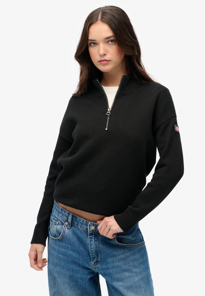 Zwarte gebreide trui met hoge kraag en halve rits; ribbelboorden en -zoom; gedragen over lichtblauwe jeans; zijkant met opvallend labeldetail.