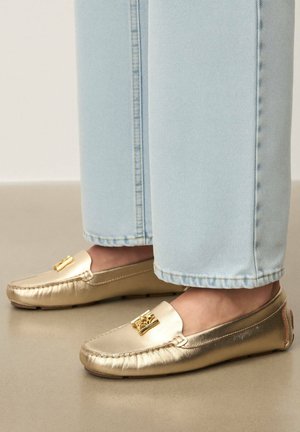 Zlate mokasinske čevlje z gladko, sijočo teksturo, z zlatim poudarkom na zgornjem delu. Kombinirane s svetlo modrimi zavihanimi kavbojkami.