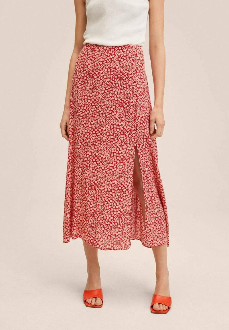 Femme portant une jupe mi-longue rouge à fleurs avec une fente avant, un haut blanc sans manches et des talons ouverts orange sur un fond uni.