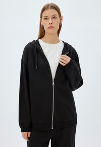 Sudadera negra con cremallera hecha de una mezcla de algodón, que presenta un corte holgado, capucha forrada, cierre de cremallera visible y puños y dobladillo acanalados.