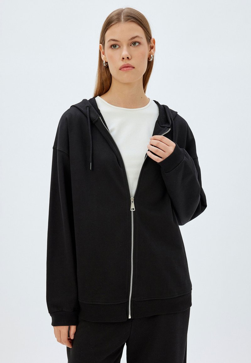 Sudadera negra con cremallera hecha de una mezcla de algodón, que presenta un corte holgado, capucha forrada, cierre de cremallera visible y puños y dobladillo acanalados.
