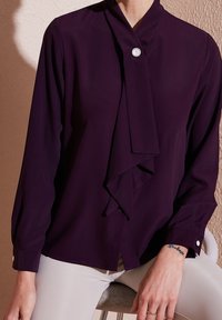 Personne portant un chemisier à manches longues violet foncé avec un bouton décoratif en perle et un pantalon clair, assise sur un tabouret.